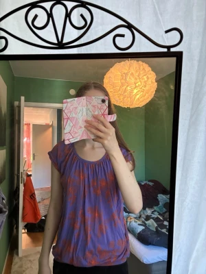 Lila blommig blus med orange mönster - Snygg lila blus med orange blommigt mönster och rund halsringning. Modellen är lös och har korta ärmar med lätt puff. Perfekt för dig som gillar färg och vill sticka ut lite extra. Tillverkad i mjuk lyocell och elastan som känns skön mot huden.