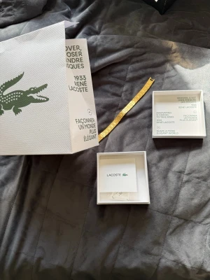 Guldfärgat armband från Lacoste - Stilrent armband från Lacoste i guldfärgad metall med fyrkantiga länkar och diskret mönstrade detaljer. Kommer i originalförpackning med både ask och tygpåse med Lacoste-logga. Perfekt accessoar för att lyfta din outfit.