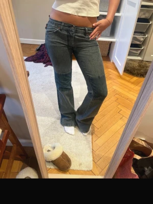 Lågmidjade bootcut jeans från michiko koshino - Säljer ett par blå vintage lågmidjade bootcut jeans från Michiko Koshino med ikonisk målad logga på bakfickorna. Jeansen har lågmidja och är gjorda i kraftigt denimtyg. Perfekta för dig som vill ha en trendig look.