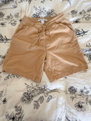 Beige shorts från Calvin Klein Jeans - Snygga beige shorts från Calvin Klein Jeans med snörning i midjan och fickor fram och bak. Mjukt bomullsmaterial och avslappnad passform, perfekta för sommaren. Diskret logga broderad på ena benet.