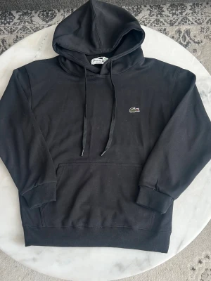Svart hoodie från Lacoste - Svart hoodie från Lacoste med klassisk krokodil-logga på bröstet. Hoodien har dragsko i huvan, magficka och ribbade muddar vid ärmslut och nederkant. Tillverkad i mjukt bomullsmaterial för en skön och avslappnad stil.