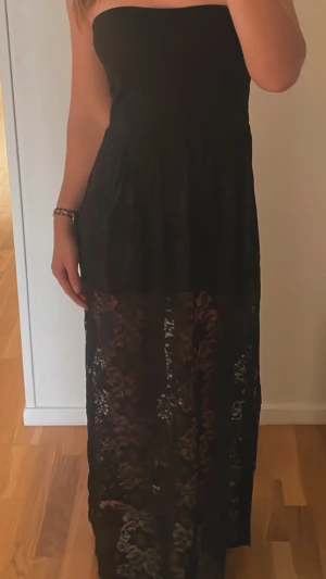 Svart axelbandslös långklänning - Snygg svart axelbandslös långklänning med transparent spetsdetalj nedtill. Klänningen har en elegant och figurnära passform, perfekt för dig som vill ha en stilren look med lite edge. Materialet känns mjukt och följsamt mot kroppen.