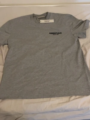 Grå Essentials Fear of God t-shirt - Snygg grå t-shirt från Essentials Fear of God med svart logga tryckt på bröstet och ryggen. Klassisk passform med rund hals och korta ärmar. Perfekt för en clean och stilren look.