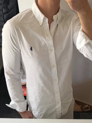 Ralph lauren skjorta - Klassisk vit skjorta från Polo Ralph Lauren med button-down krage och broderad logga på bröstet. Skjortan har lång ärm, knappar framtill och är tillverkad i bomull. Perfekt för en clean och stilren look.