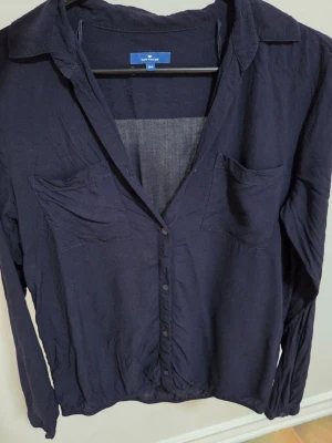 Mörkblå skjorta/blus från Tom Tailor - Snygg mörkblå blus från Tom Tailor med klassisk krage, två bröstfickor och knappar framtill. Blusen är långärmad och tillverkad i viskos som håller dig sval, perfekt för en avslappnad men stilren look.