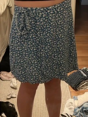 Blå blommig omlottkjol med knyt - Söt blå omlottkjol med vitt blommönster och knyt i midjan. Kjolen har en kort längd och är perfekt för varma dagar. Materialet känns lätt och luftigt, vilket gör den skön att bära. Passar dig som gillar en avslappnad men ändå söt stil.