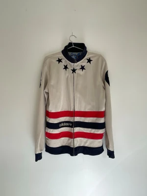 Adidas vindjacka med stjärnor och ränder - Snygg vindjacka från Adidas i beige med marinblå och röda ränder samt stjärnor runt kragen. Jackan har hel dragkedja, muddar i ärmslut och nederkant samt Adidas-logga framtill. Perfekt för en sportig och retro look.