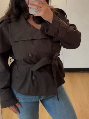 Kort brun trenchcoat med knytskärp - Säljer en helt ny brun trenchjacka/kavaj från Bershka￼ 🤎 Fortfarande kvar prislapp på!  Superfin modell med knytning i midjan som ger en snygg och stilren passform. Perfekt till både vardag och finare outfits nu till våren/hösten. Trendig mörkbrun färg och jättefin på. 