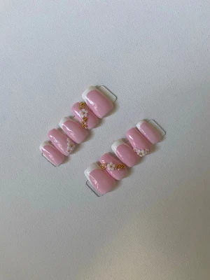 Rosa press-on naglar med blommor - Snygga press-on naglar i ljusrosa med vit fransk tipp och dekorerade med små vita blommor och guldglitter. Naglarna har en oval form och ger en söt och trendig look. Perfekt för dig som vill ha en feminin och stilren manikyr på nolltid.