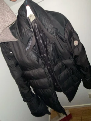 Svart dunjacka från Moncler - Snygg svart dunjacka från Moncler med klassisk puffer-design och märkets logga på ärmen. Jackan har hög krage, dragkedja och är fodrad med certifierat dun enligt DIST. Perfekt för kalla dagar och ger en stilren look.