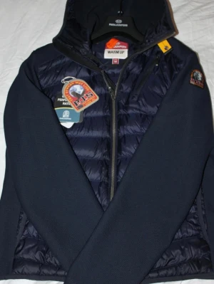Mörkblå hybridjacka från Parajumpers - Snygg mörkblå hybridjacka från Parajumpers med vadderad front och rygg i glansigt material samt stickade ärmar. Jackan har huva, dragkedja och patch på ärmen. Perfekt för dig som vill ha en sportig och trendig look.