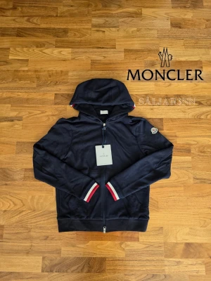 Marinblå Moncler Hoodie - Moncler Hoodie – Marinblå | Storlek S | Nyskick Exklusiv Moncler-hoodie i marinblå färg, storlek S, helt ny. Perfekt för vardag eller stilfull streetwear. Hoodien har dragkedja med Moncler-logga, ribbstickade ärmslut med röda, vita och blå detaljer, samt Moncler-emblem på vänster ärm. Mjuk, bekväm passform med huva gör den både funktionell och trendig. 📦 Snabb leverans – skickas samma dag (om ej hindr.) 📩 Kontakta vid frågor.