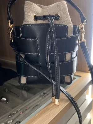 Svart och beige bucket bag med axelrem - Trendig bucket bag med svart läder och vita kontrastsömmar, kombinerat med beige canvas. Väskan har justerbar axelrem, guldfärgade metalldetaljer och dragskostängning. Perfekt för dig som gillar stilrena och moderna accessoarer.