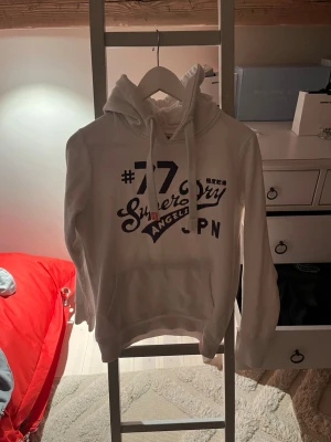Vit hoodie från Superdry med tryck - Tja! Säljer en vit hoodie från Superdry med stort tryck på bröstet i svart och lila, samt texten 'Superdry Angels JPN'. Klassisk modell med huva och dragsko, samt känguruficka framtill. Perfekt för en avslappnad och sportig stil enligt mig. Riktigt bra skick och Säljer för att den blivit för liten. Storlek s men skulle säga att den passar dig som har xs med längden 170-180. Skriv om ni har funderingar och pris går att diskutera 