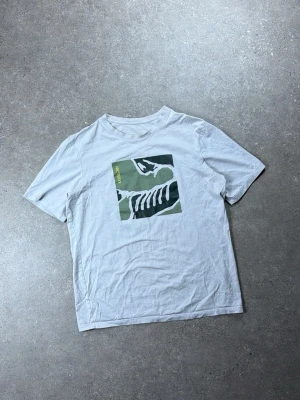 Vit Arc'teryx t-shirt med tryck - Vit t-shirt från Arc'teryx med ett stort grafiskt tryck i grönt och svart på bröstet. Klassisk rund hals och korta ärmar. Perfekt för en avslappnad och cool stil.