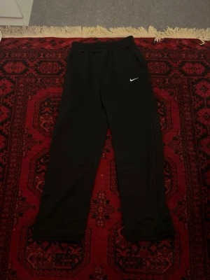 Svarta mjukisbyxor från Nike - Snygga svarta mjukisbyxor från Nike med klassisk vit logga på vänster ben. Byxorna har resår i midjan och sidofickor, perfekta för chill eller träning. Tillverkade i mjukt material som känns skönt mot huden. Enkel och stilren design som funkar till det mesta.