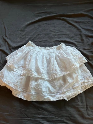 Vit volangkjol med spetsdetaljer - Säljer en vit kortkjol med flera volanger och fina spetsdetaljer. Kjolen har elastisk midja och är gjord i ett lätt bomullsmaterial som känns skönt mot huden. Perfekt för dig som gillar romantiska och luftiga plagg.