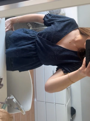 Mörkblå omlottblus med spetsdetaljer - Snygg mörkblå omlottblus med korta ärmar och fina spetsdetaljer. Blusen har v-ringning och volangkant vid ärmslut och nederkant. Perfekt att styla med jeans eller kjol för en trendig look.