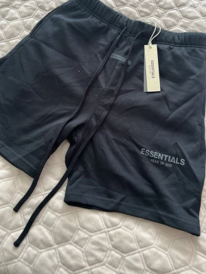 Svarta shorts från Essentials Fear of God - Svarta shorts från Essentials Fear of God med tryckt logga på vänster ben. Modellen har elastisk midja med snörning och är tillverkad i mjuk bomull. Perfekta för en avslappnad och trendig stil.