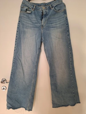 Ljusblåa raka jeans med rå kant - Säljer ett par ljusblå jeans med rak passform och rå, fransig kant nertill. Jeansen har klassisk femficksdesign och hög midja. Perfekta för en avslappnad och trendig look. Materialet är denim i bomull.