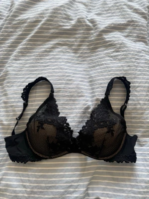 Svart intimissimi bh - Snygg svart bh med spetsdetaljer och vadderade kupor. Justerbara axelband med spets och hakknäppning bak. Kuporna har ett diskret rutmönster och små rosetter framtill. Perfekt för dig som vill ha en feminin och bekväm bh.