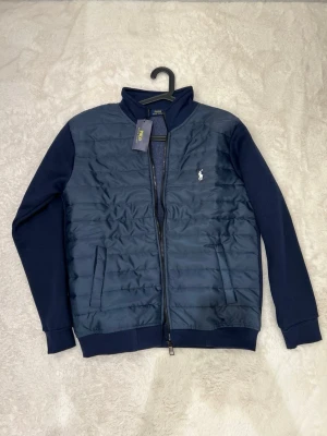 Marinblå cardiganjacka Polo Ralph Lauren - Snygg marinblå cardiganjacka från Polo Ralph Lauren med quiltad framsida, dragkedja och två fickor. Jackan har ribbade muddar och klassisk logga broderad på bröstet. Perfekt för lager-på-lager och stilrena outfits.