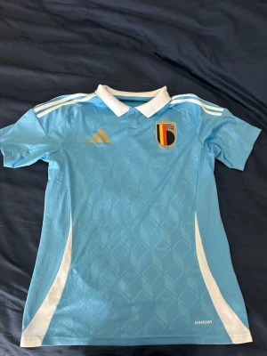 Belgien Adidas fotbollströja blå - Snygg ljusblå Belgien fotbollströja från Adidas med vita detaljer och klassisk krage. Tröjan har Belgien-emblemet på bröstet, tre vita ränder på axlarna och diskret mönster i tyget. Tillverkad i lätt och ventilerande material med AEROREADY-teknologi.