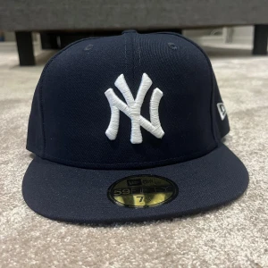 New York Yankees keps från New Era 6 7/8 - Snygg marinblå New York Yankees keps från New Era, modell 59FIFTY. Kepsen har en rak skärm och broderad vit NY-logga framtill samt MLB-logga baktill. Tillverkad i polyester för en skön och slitstark känsla. Perfekt för dig som gillar streetstyle och sport. OBS KEPSEN ÄR HELT SPRILLANS NY OCH OANVÄND.