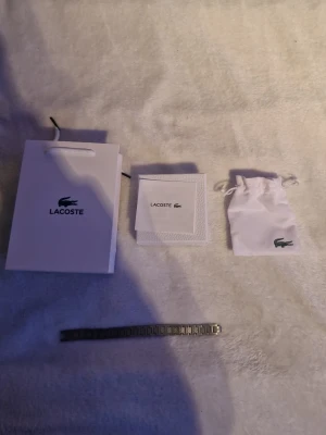 Silverfärgat armband från Lacoste - Stilren silverfärgat armband från Lacoste med länkarmband i metall och klassisk krokodil-logga på spännet. Kommer med originalpåse, ask och påse från Lacoste. Perfekt accessoar för dig som gillar en clean och tidlös look.