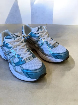 Asics gel-1130 vita och blå sneakers - Fräscha sneakers från Asics i vitt meshmaterial med blå och silverfärgade detaljer. Skorna har klassisk låg profil, vita skosnören och en bekväm sula som passar perfekt till en sportig stil. Snygga kontraster mellan färgerna och ikoniska Asics-loggan på sidan.