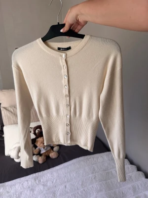 Krämvit kofta från Gina Tricot - Snygg krämvit kofta från Gina Tricot med rund halsringning och pärlemorknappar framtill. Långärmad modell med ribbade muddar och midjekort passform. Perfekt att styla över en topp eller klänning.