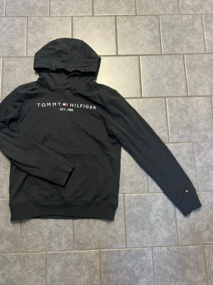 Svart hoodie från Tommy Hilfiger - Svart hoodie från Tommy Hilfiger med vit logga och flagga på bröstet samt liten logga på ärmen. Klassisk känguruficka framtill och ribbade muddar. Perfekt för en avslappnad och stilren look.