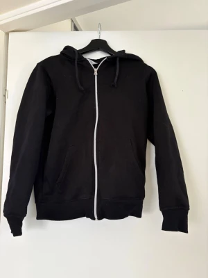 Svart zip-hoodie med huva - En klassisk svart hoodie med dragkedja framtill och två fickor. Modellen har huva med dragsko och ribbade muddar vid ärmslut och nederkant. Perfekt för en chill och avslappnad stil. Materialet känns mjukt och bekvämt, troligen bomullsblandning. Lite nopprig inuti 