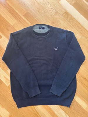 Mörkblå tröja från Gant - Mörkblå långärmad tröja från Gant med rund halsringning och diskret logga broderad på bröstet. Tröjan har ribbade muddar vid ärmslut och nederkant. Perfekt för en stilren och klassisk look.