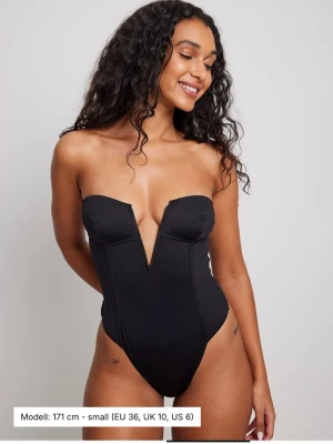 Svart bandeau body med djup V-ringning - Snygg svart body ifrån NAKD, perfekt till fest, stylat med en kavaj, jeans eller kostymbyxor. Jätteskönt material och väldigt stretchig. Den är helt oanvänd då jag inte får anständig för den, den är i storlek xxs men skulle säga att den passar om man har upp till storlek s då det är min normala storlek.
