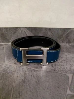 Blått bälte - Snyggt blått skärp med ett stort, silverfärgat H-spänne. Skärpet har vita sömmar längs kanterna och är gjort i slätt skinn. Perfekt för att lyfta vilken outfit som helst.