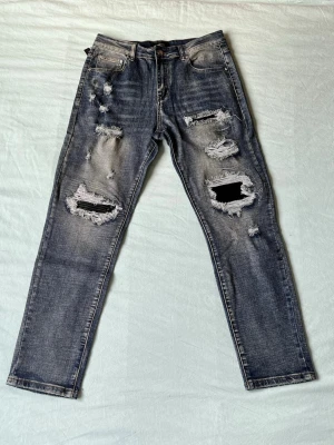 Grå slitna jeans med stora hål - Säljer ett par grå jeans med rejält slitna detaljer och stora hål på båda benen. Jeansen har en rak passform och klassisk femficksdesign. Perfekta för en edgy streetstyle-look. Materialet är denim i bomull.