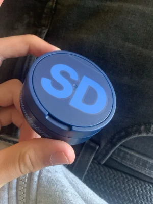 Blå snusdosa med SD-logga - En blå snusdosa i plast med SD-logga på locket och texten 'Sverigedemokraterna' på sidan. Dosan har ett gångjärnslock och är kompakt och rund i formen. Perfekt för att förvara snus eller småsaker.