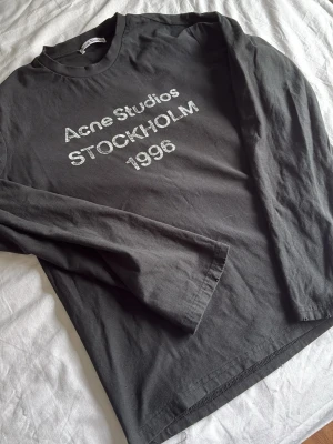 Svart långärmad topp Acne Studios - Svart långärmad topp från Acne Studios med tryck 'Acne Studios STOCKHOLM 1996' framtill. Klassisk rund halsringning och rak passform. Perfekt för dig som gillar stilrena och trendiga plagg med tydlig logga.