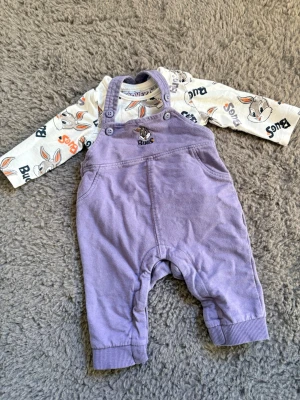Looney Tunes babyset med lila hängselbyxa - Sött babyset med lila hängselbyxa i mjuk bomull och långärmad body med Bugs Bunny-tryck. Hängselbyxan har broderad Bugs Bunny och knappar på axelbanden. Perfekt för små fans av Looney Tunes. Den är använd 2 ggr 