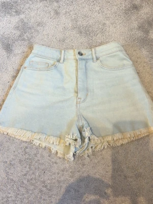 Ljusblåa högmidjade jeansshorts - Säljer ett par ljusblåa högmidjade jeansshorts med fransig kant och klassisk femficksdesign. Shortsen har dragkedja och knapp framtill samt bälteshällor. Perfekta för sommaren och ger en avslappnad vibe.