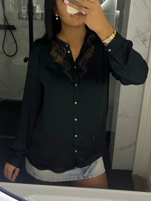 Svart blus med spetsdetaljer - Snygg svart blus med långa ärmar och knappar framtill. Blusen har v-ringning och fina spetsdetaljer vid halsringningen som ger en elegant touch. Tillverkad i ett glansigt, mjukt material som känns lyxigt.