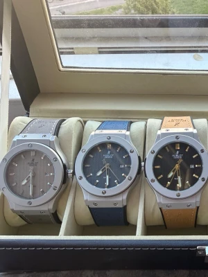   HUBBLLOOTT Classic Fusion klockor 1300 st -  Classic Fusion klockor med rund boett i silverfärgad metall och synliga skruvar. En har grå urtavla och grått band, en har blå urtavla och blått band, och en har brun urtavla med brunt band. Alla har datumvisning och stilren design.