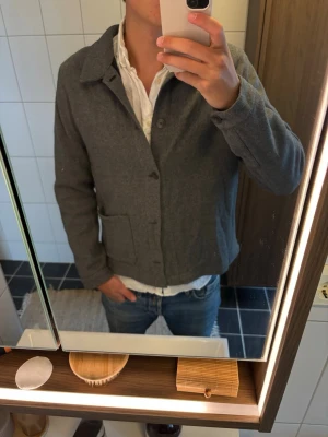 Grå ullcardigan med fickor - Stilren grå cardiganjacka i ullmix med klassisk krage och stora framfickor. Jackan har knäppning framtill och en rak passform som ger en clean look. Perfekt att slänga över en skjorta för en avslappnad men snygg stil. Två knappar av