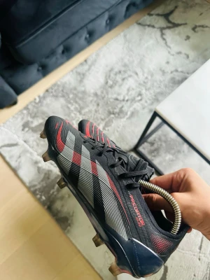 Adidas predator elite - Säljer ett par Adidas Predator fotbollsskor i svart med röda och silvriga detaljer. Skorna har klassiska tre ränder på sidan, snörning och dobbar för spel på gräs. Materialet är syntet med textur för bättre bollkontroll. Perfekta för dig som vill dominera på planen.