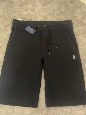 Svarta shorts från Polo Ralph Lauren - Svarta shorts från Polo Ralph Lauren med klassisk logga broderad i vitt på benet. Modellen har snörning i midjan och två sidofickor. Perfekta för en avslappnad och stilren look.