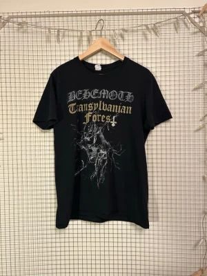 Behemoth Transylvanian Forest t-shirt - Säljer en ovanlig svart band-tisha från Behemoth med tryck fram och bak. På framsidan står det 'Transylvanian Forest' i gotisk stil och har en mörk grafisk illustration. Baksidan har bandets logga och bild på medlemmarna. Perfekt för dig som gillar metal och vill sticka ut.