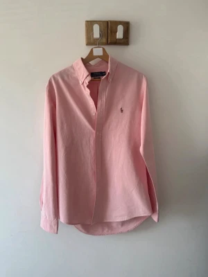 Rosa skjorta från Polo Ralph Lauren - Klassisk rosa skjorta från Polo Ralph Lauren med button-down krage och broderad logga på bröstet. Skjortan har lång ärm och knappar framtill. Perfekt för en stilren och avslappnad look.