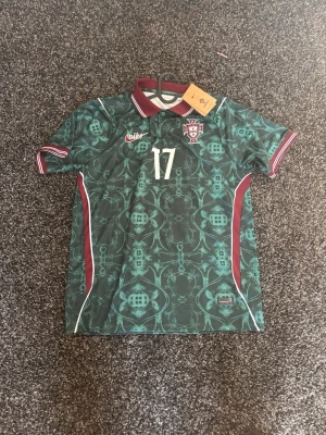 Portugal fotbollströja R. LEÃO 17 - Snygg Portugal fotbollströja från Nike med nummer 17 och namnet R. Leão på ryggen. Tröjan är grön med mönster, vinröda detaljer och vit logga samt tryck. Klassisk krage med röd och vit rand, korta ärmar och sportig passform. Perfekt för match eller träning.