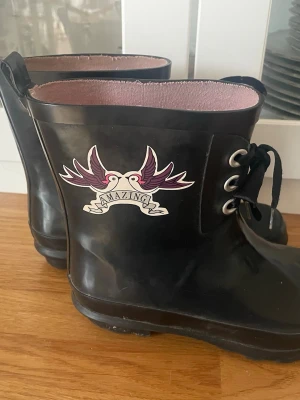 Svarta gummistövlar med tryck - Säljer ett par svarta gummistövlar med rosa innerfoder och coolt tryck med två fåglar och texten 'Amazing' på utsidan. Stövlarna har snörning framtill och dragflik bak för enkel påtagning. Perfekta för regniga dagar och festivaler.